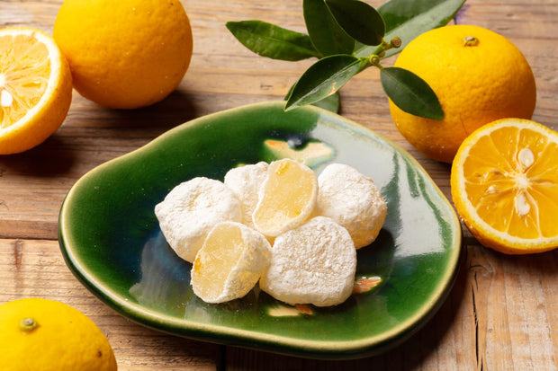 Six powdered mochi with yellow citrus filling are arranged on a green ceramic plate, surrounded by whole and halved yellow citrus fruits with leaves, all atop a wooden surface—an inviting scene that captures the spirit of Japanese snacking and echoes the vibrant assortment found in the Dagashi Snack Crate - 30-Piece Japanese Snack Variety Box with Candy, Chocolate, Cookies, Mochi & Ramune Treats.