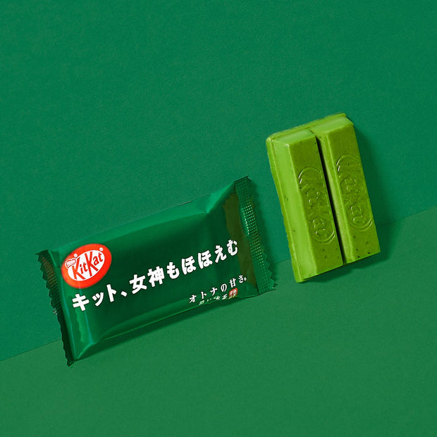 A green-wrapped KitKat bar with Japanese text is displayed beside two unwrapped green KitKat pieces on a vibrant green background, offering a tempting preview of the authentic treats featured in the Dagashi Snack Crate—your 30-piece Japanese Snack Variety Box packed with candy, chocolate, cookies, mochi, and Ramune delights.