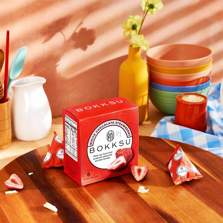 A Bokksu White Strawberry 6-Piece Box (2-Pack) sits on a wooden surface, encircled by ceramic bowls, a vase, and cutlery. These Japanese artisanal snacks offer chocolate-infused strawberries for an indulgent treat. thumbnail 2