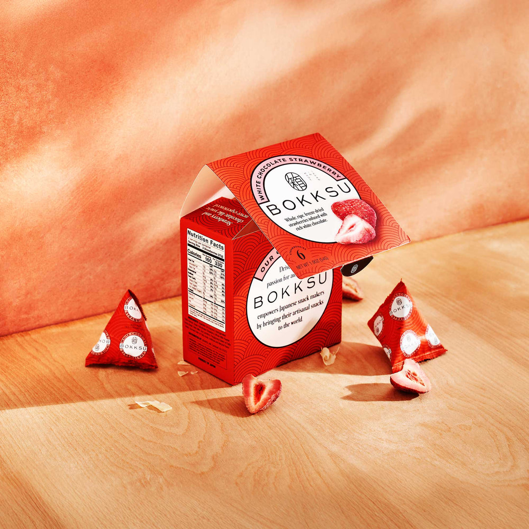 A vibrant red box of Bokksu White Strawberry 6-Piece Box (2-Pack), displaying opened packets and an array of scattered strawberries, sits invitingly on a wooden surface against an orange backdrop. This irresistible Japanese treat from Bokksu masterfully blends the tart flavor of fresh strawberries with the creamy sweetness of white chocolate.