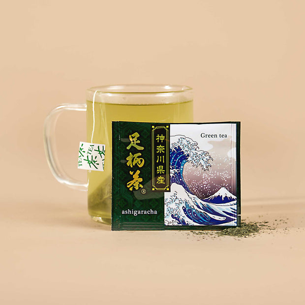 A glass mug of green tea sits behind The Japanese Tea Box sachet, which features a wave illustration, with matcha powder scattered on the surface.