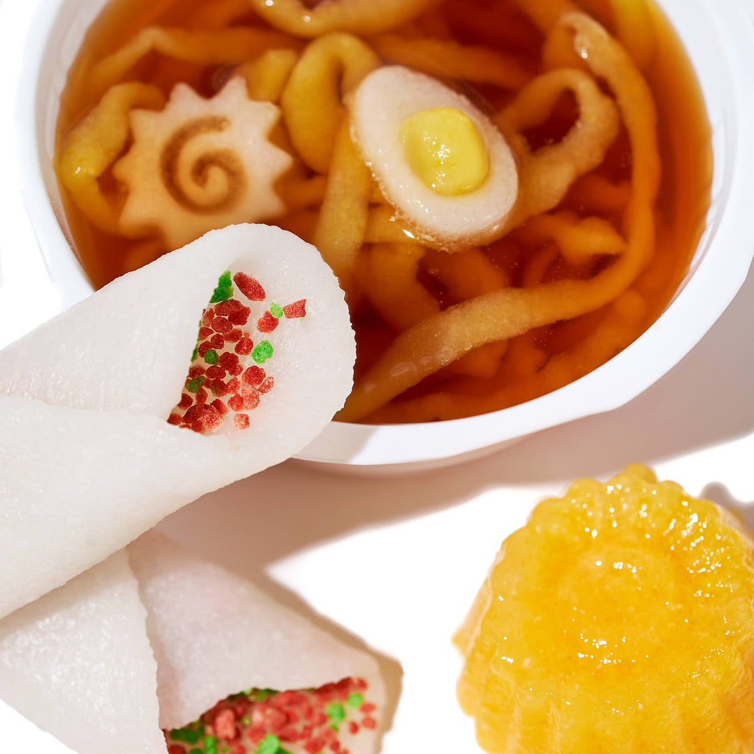 A bowl of soup adorned with decorative food items is paired with two rolled treats topped with vibrant red and green accents, reminiscent of the Bokksu Market DIY Candy Kit Pack (6-Pack), along with a yellow, flower-shaped dessert on a white background, all artfully crafted to mimic Kracie's precision in homemade candies.