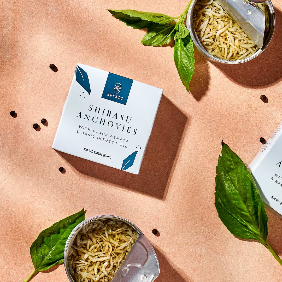 Two opened cans of Bokksu Tinned Shirasu Anchovies with Black Pepper and Basil-Infused Oil (2-Pack) rest on a peach-colored surface, surrounded by fresh basil leaves. Packaging boxes emblazoned with the Bokksu brand are also visible, evoking the perfect starter for an umami spaghetti dish.