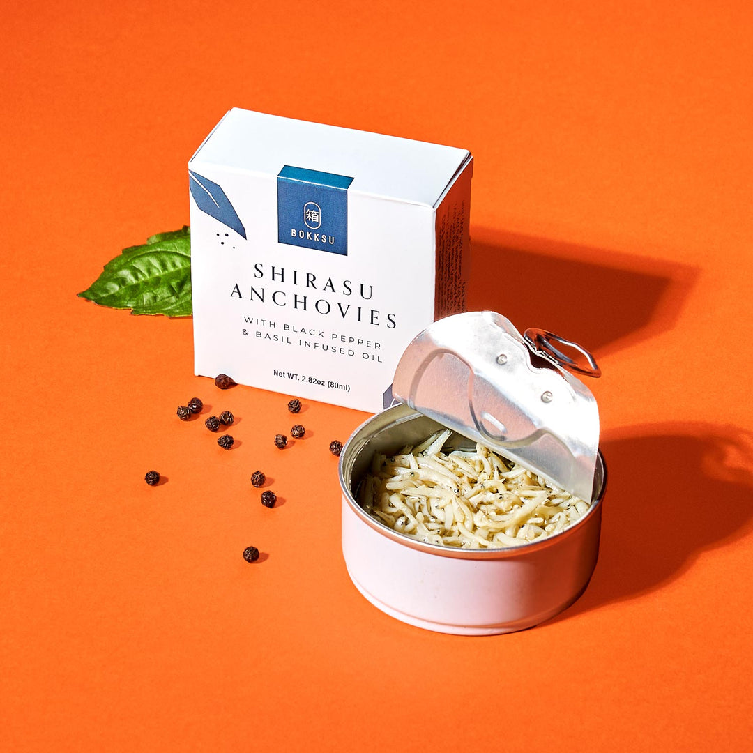 A tin of Bokksu Tinned Shirasu Anchovies with Black Pepper and Basil-Infused Oil (2-Pack), next to its box, on an orange background with scattered peppercorns and a basil leaf. Perfect for adding an umami twist to your favorite spaghetti dish.