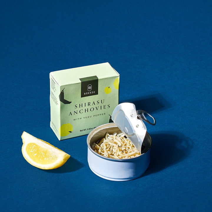 A box of "Bokksu Tinned Shirasu Anchovies with Yuzu Pepper," known for being high in protein, is displayed alongside an opened tin of anchovies and a lemon wedge on a blue surface.