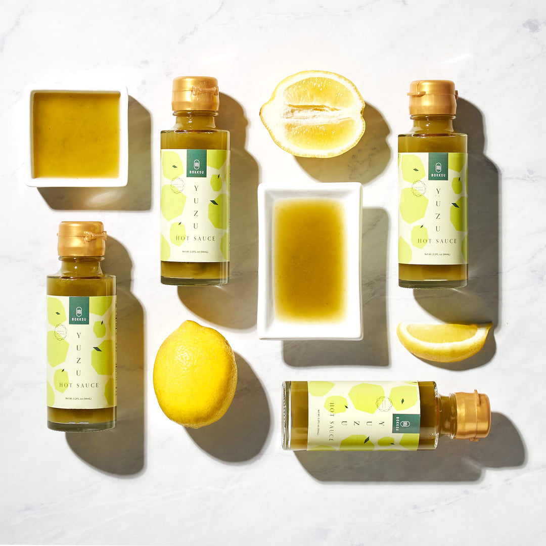 Overhead view of four bottles of Bokksu Yuzu Hot Sauce, a perfect Tobasco alternative, arranged on a light surface with whole and halved lemons, and small containers of greenish liquid.