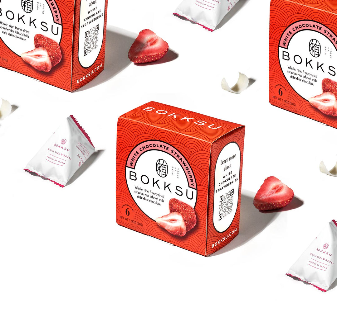 A Bokksu White Strawberry 6-Piece Box (2-Pack) by Bokksu is displayed alongside individually packaged pieces of this artisanal Japanese snack and strawberry slices on a white background.