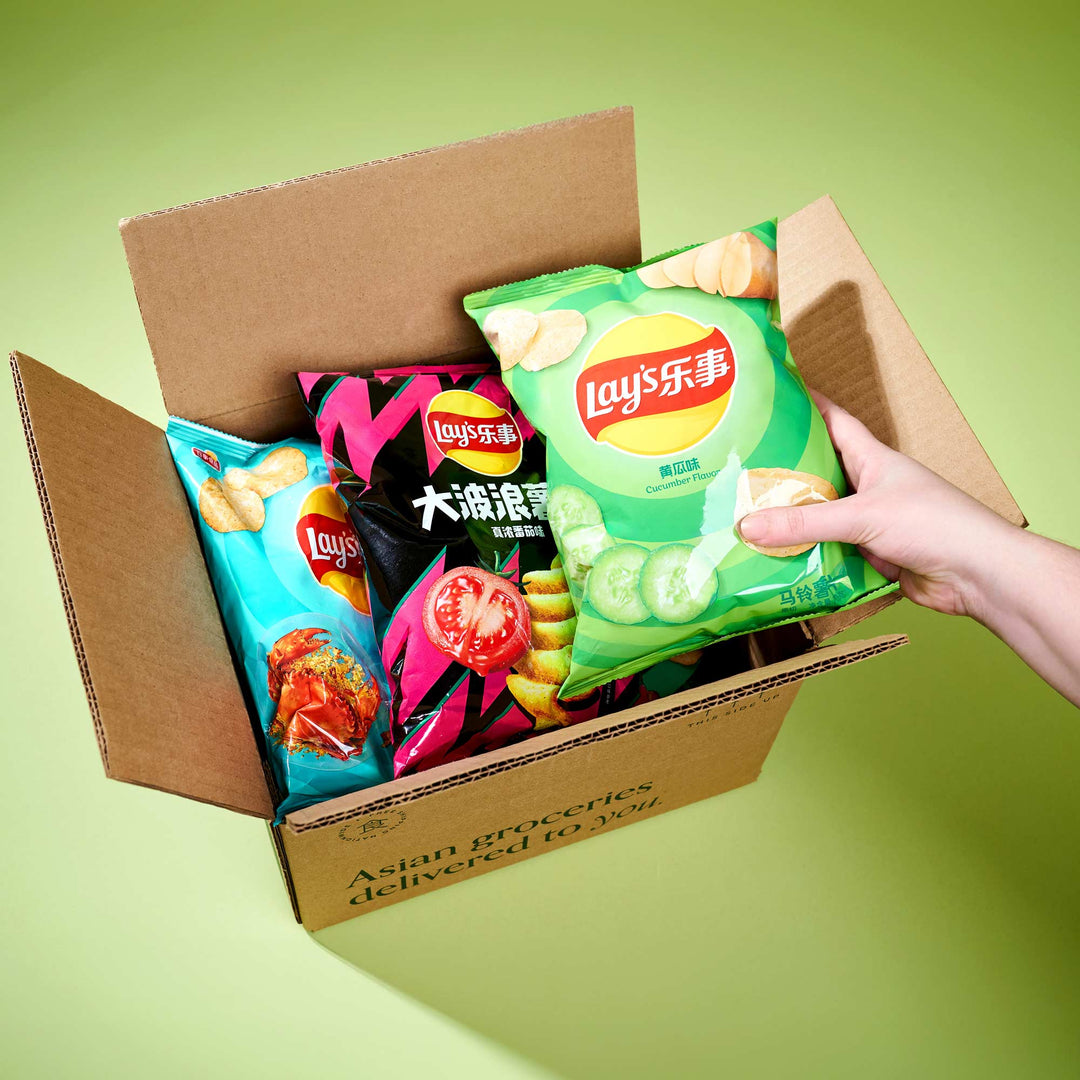 A hand holds a bag of cucumber-flavored Lay's chips from the Bokksu Market Discovery Pack over an open box, which contains two more bags of Lay's in different flavors, offering a delightful experience of Asian snacks.