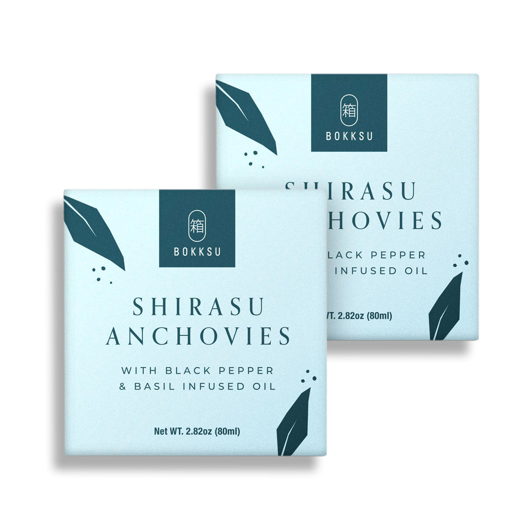 Two teal-colored boxes of Bokksu Tinned Shirasu Anchovies with Black Pepper and Basil-Infused Oil (2-Pack). Each box weighs 2.82oz (80ml). Perfect for crafting a delectable Umami Spaghetti dish.