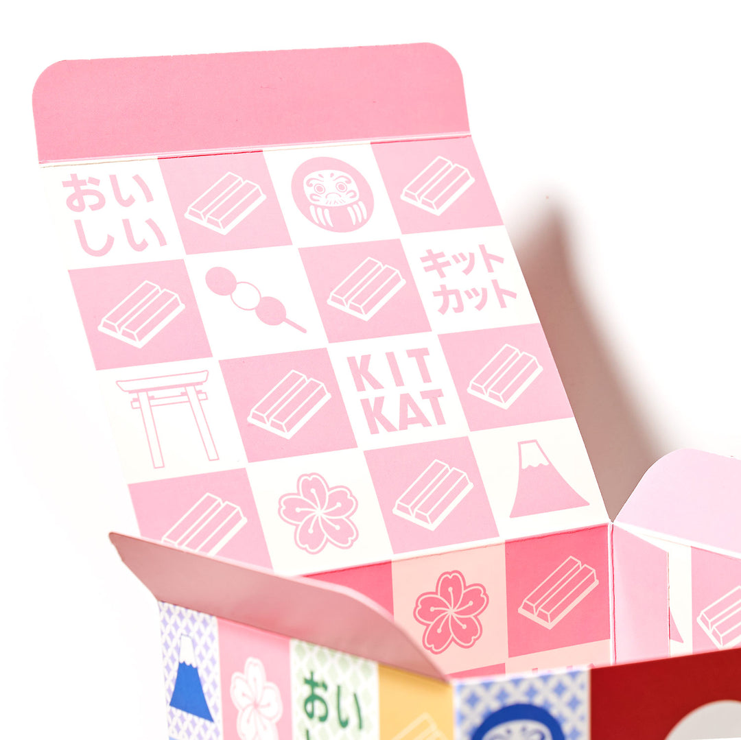 Open the Kit Kat Sampler (50 pcs.) box from Bokksu Market adorned with pink-themed Japanese text and illustrations depicting Kit Kat bars, a torii gate, a Daruma doll, Mount Fuji, and various other symbols. This variety pack celebrates the essence of Japanese Kit Kat culture with an assortment of unique flavors.