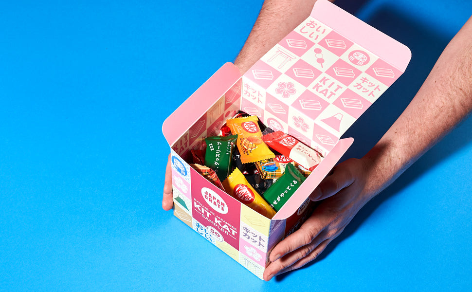 Hands holding an open colorful box containing the Kit Kat Sampler (50 pcs.) by Bokksu Market, featuring an assortment of unique Japanese Kit Kat flavors and assorted Japanese candies, displayed against a blue background.