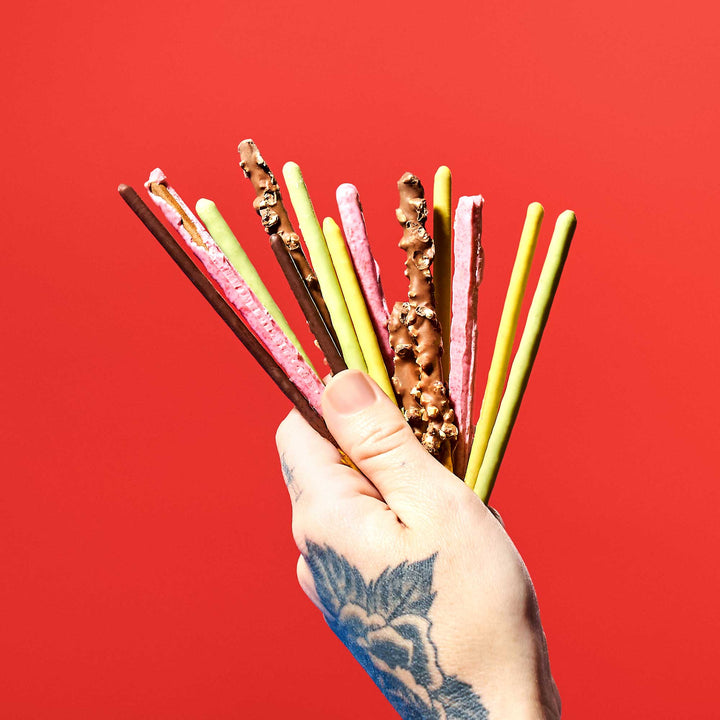 A hand adorned with a floral tattoo holds a variety pack of Glico Pocky biscuit sticks against a vibrant red background, each offering a distinct flavored chocolate coating. thumbnail 2