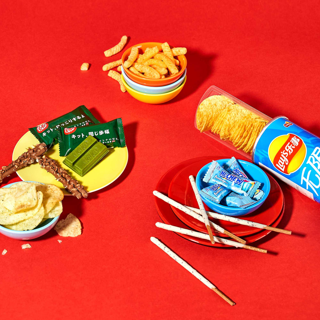 Introducing the Sweet & Savory Snack Mix by Bokksu Market, a delicious assortment featuring chips, KitKat bars, Pocky sticks, cheese curls, and gum, all elegantly displayed on a vibrant red background.