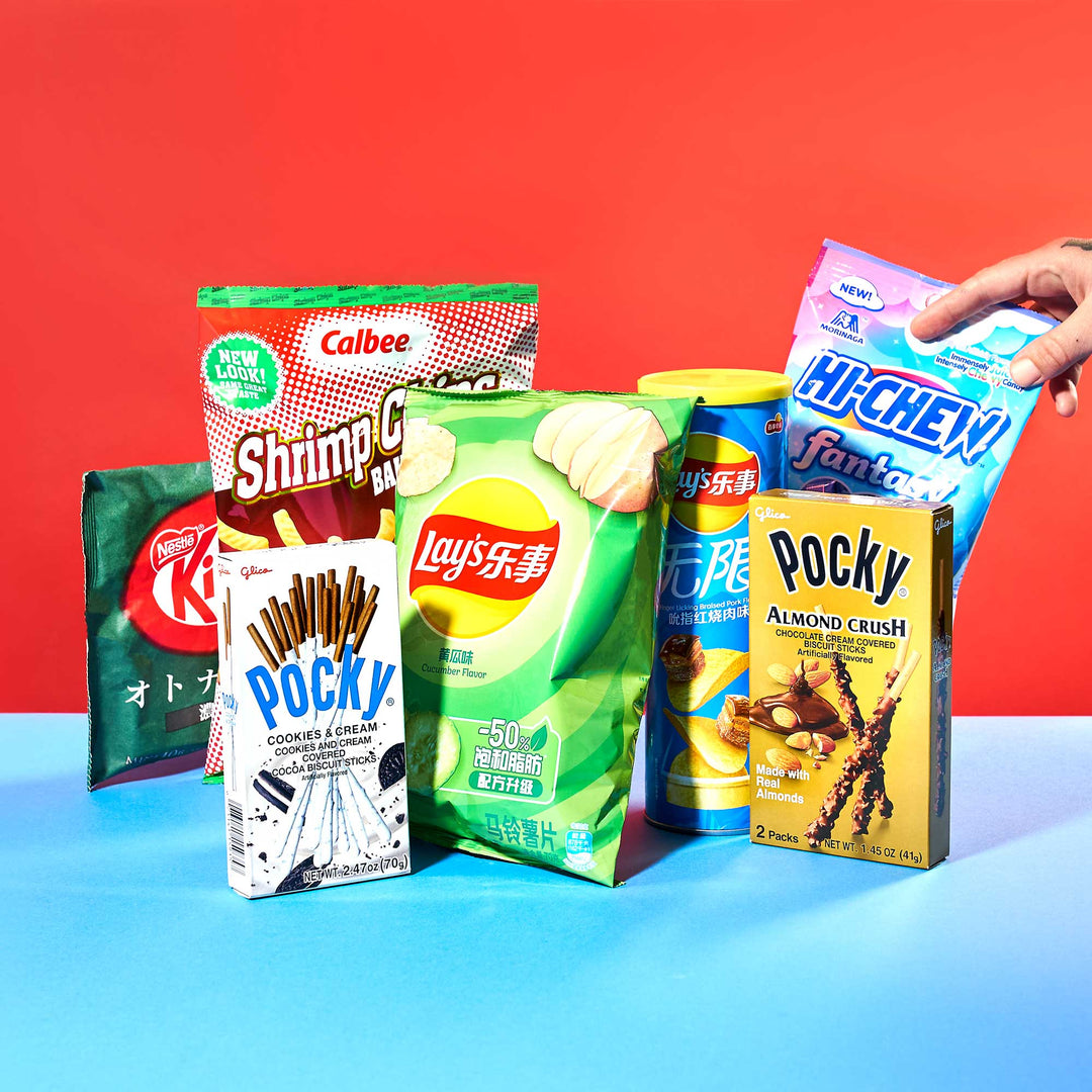 A hand reaches for the Sweet & Savory Snack Mix by Bokksu Market, showcasing an assortment of Asian favorites like Pocky, Lay's, KitKat, and Hi-Chew, beautifully arranged on a blue surface.