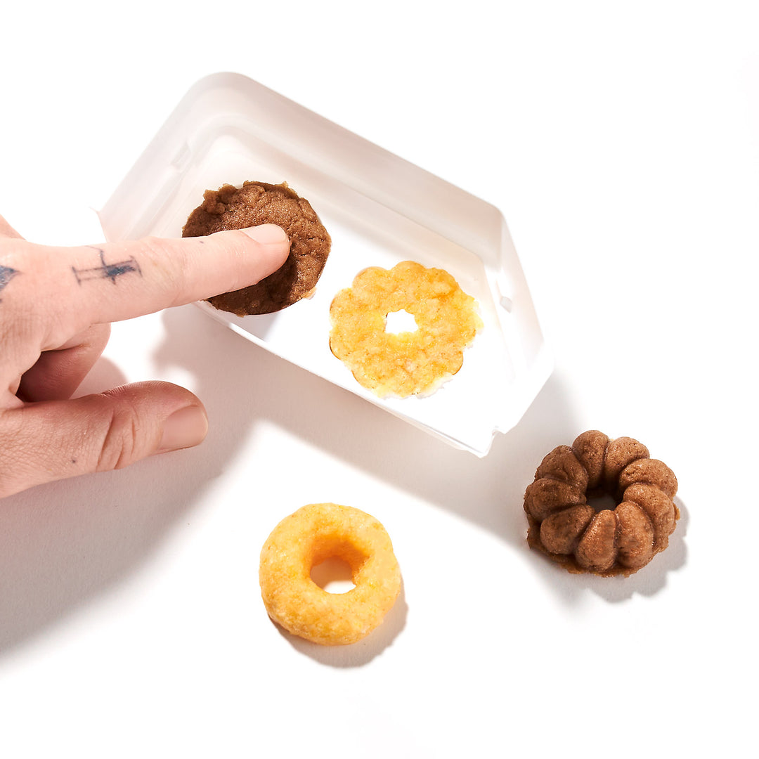 A hand reaches for a brown doughnut-shaped dog treat in a white container, reminiscent of the perfectly crafted designs from the DIY Candy Kit Pack by Bokksu Market. Three additional treats rest outside on the pristine white surface.