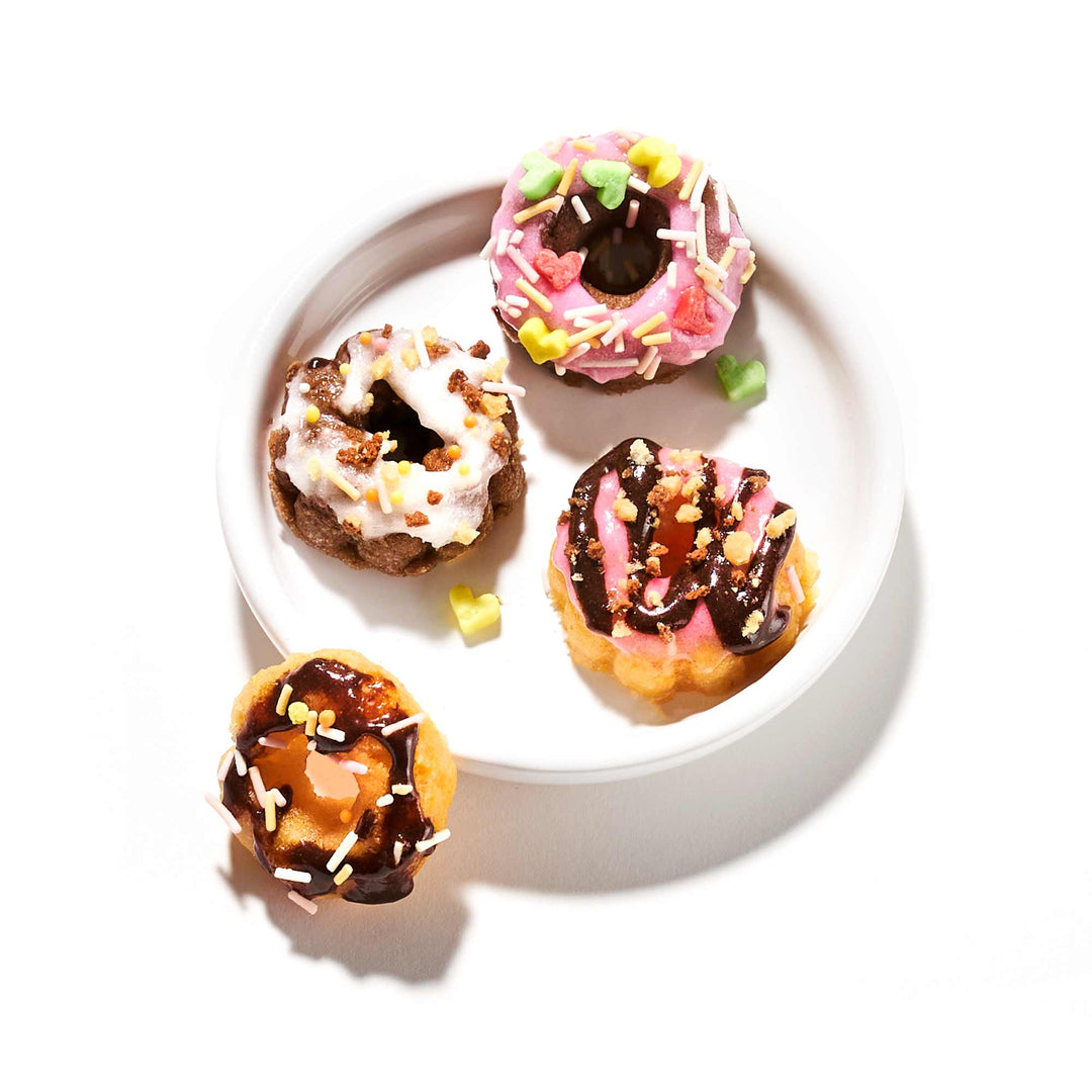 A delightful assortment of four miniature donuts, each embellished with various icings and sprinkles, is beautifully presented on a white plate, with one donut placed beside it. Create these enjoyable treats using the DIY Candy Kit Pack (6-Pack) from Bokksu Market for a fun homemade candy-making experience that captures the joy of Kracie creations.