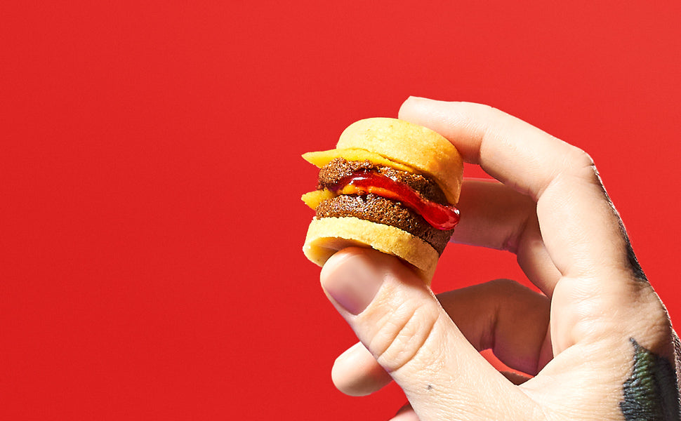 Close-up of a hand holding a small slider burger with a patty, cheese, and a dollop of ketchup against a solid red background, reminiscent of a Kracie Popin Cookin DIY Candy: Hamburger from Kracie.