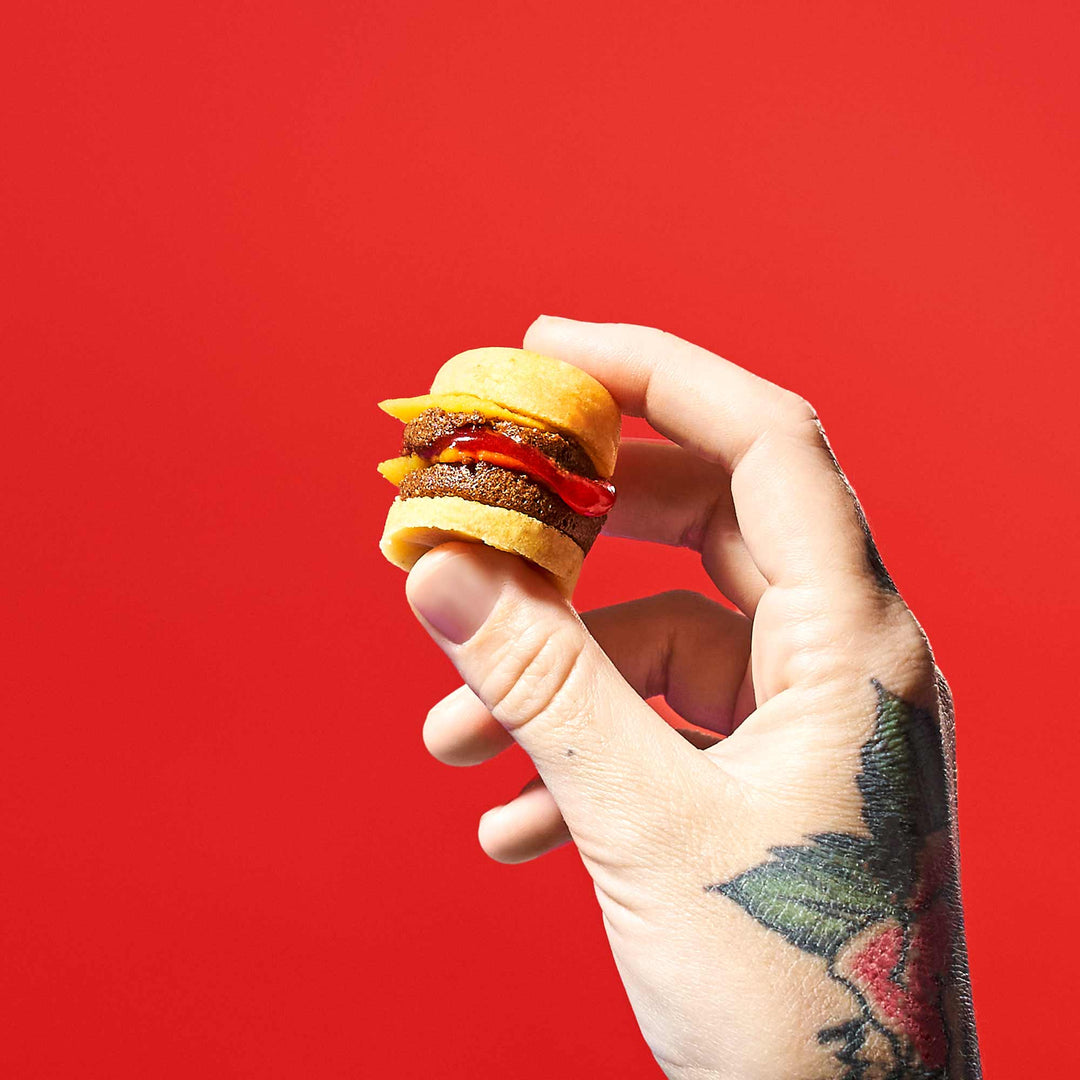 A tattooed hand holds a miniature cheeseburger, reminiscent of the Bokksu Market DIY Candy Kit Pack (6-Pack), set against a bright red background.