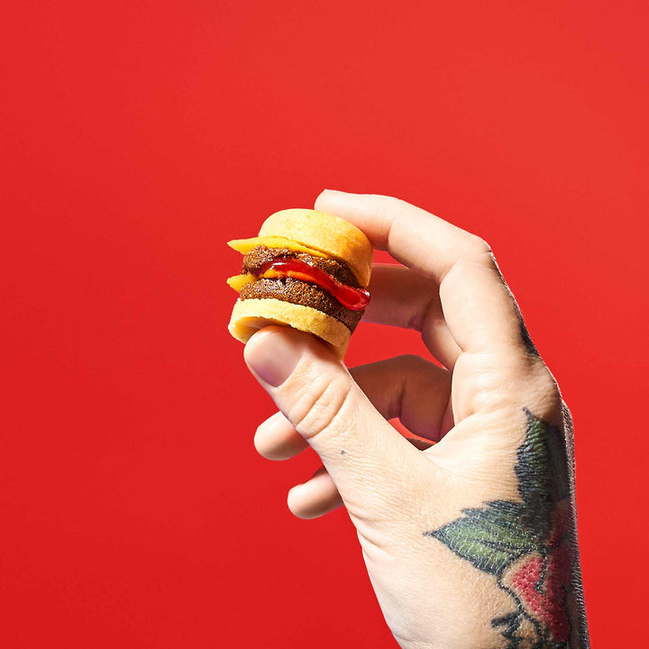 A tattooed hand holds a miniature cheeseburger, reminiscent of the Bokksu Market DIY Candy Kit Pack (6-Pack), set against a bright red background. thumbnail 4