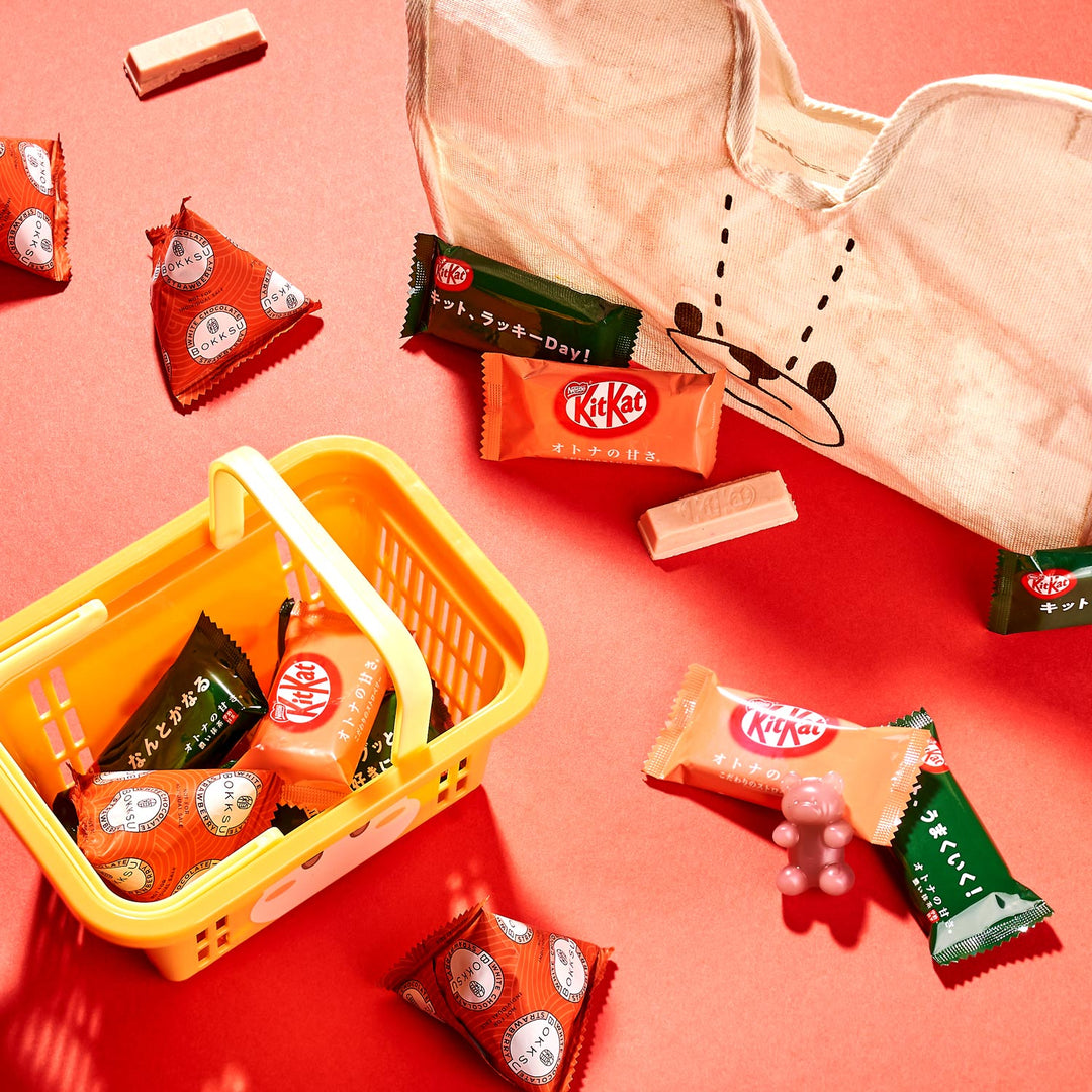 Assorted KitKat bars in orange, green, and red wrappers are spread on a red surface, accompanied by a cloth bag and a small yellow basket from the Bokksu Kawaii Gift Pack - Stationery Surprises & Japanese Chocolate Treats.