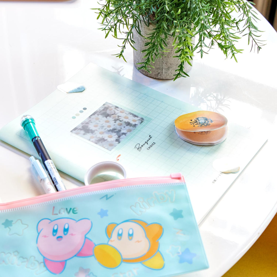 A white table is adorned with the "Kawaii Gift Pack - Stationery Surprises & Japanese Chocolate Treats" by Bokksu, featuring notebooks, pens, pencils, erasers, and a pouch with cartoon characters. A potted plant adds natural charm to the scene.