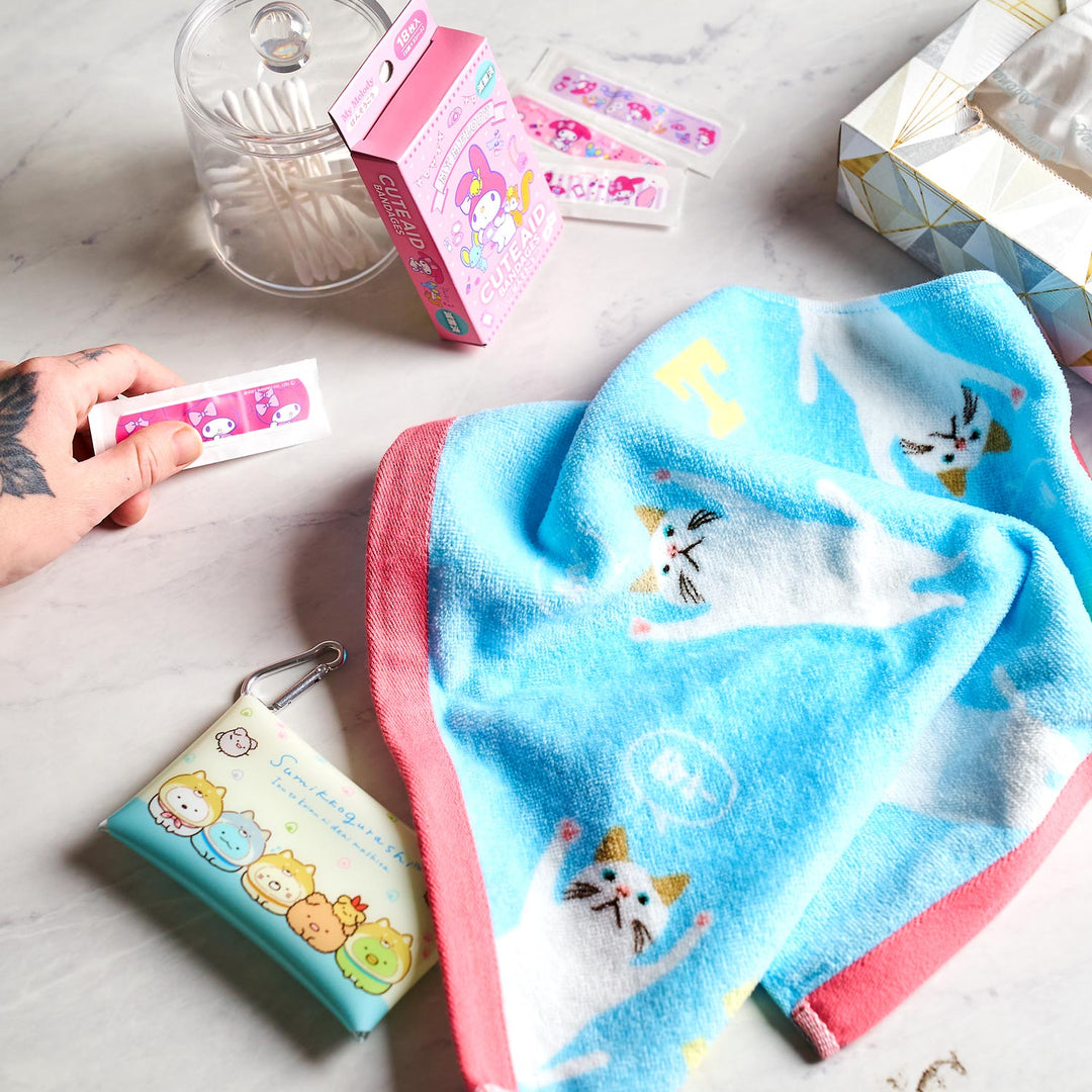 A hand holds a pink bandage near Bokksu's Holiday Gift Pack, featuring Japan's cutest items like a decorative tin, a box of bandages, tissues, and a blue towel with cat designs arranged on the table.