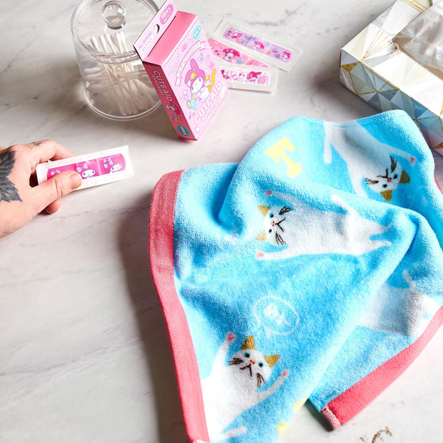 A hand holds a pink bandage near cute bandages, a blue cat towel, tissue box, and clear container—perfect for pairing with your Kawaii Gift Pack - Stationery Surprises & Japanese Chocolate Treats on a marble surface.