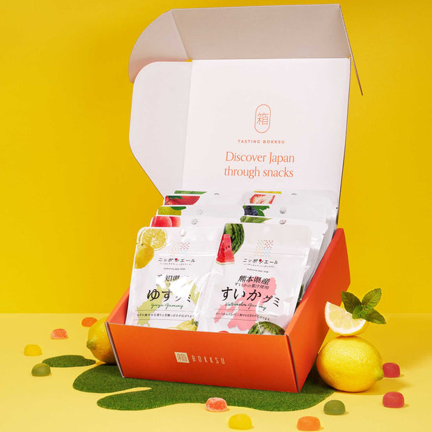An open Japanese Fruits Gummy Box displays a colorful assortment of fruit gummies, with additional gummies and whole lemons scattered around on a vibrant yellow background.