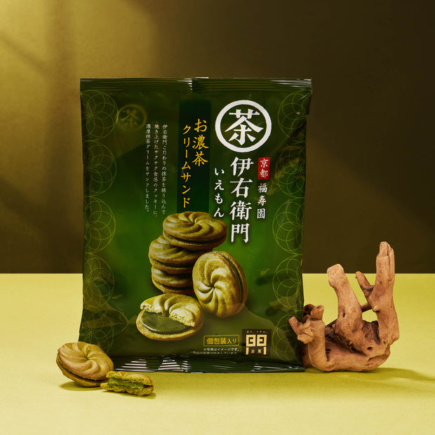The Matcha Moments Box—a green package of Japanese matcha cream sandwich cookies—stands upright beside decorative wood on a yellow surface and background, making it a perfect matcha gift.