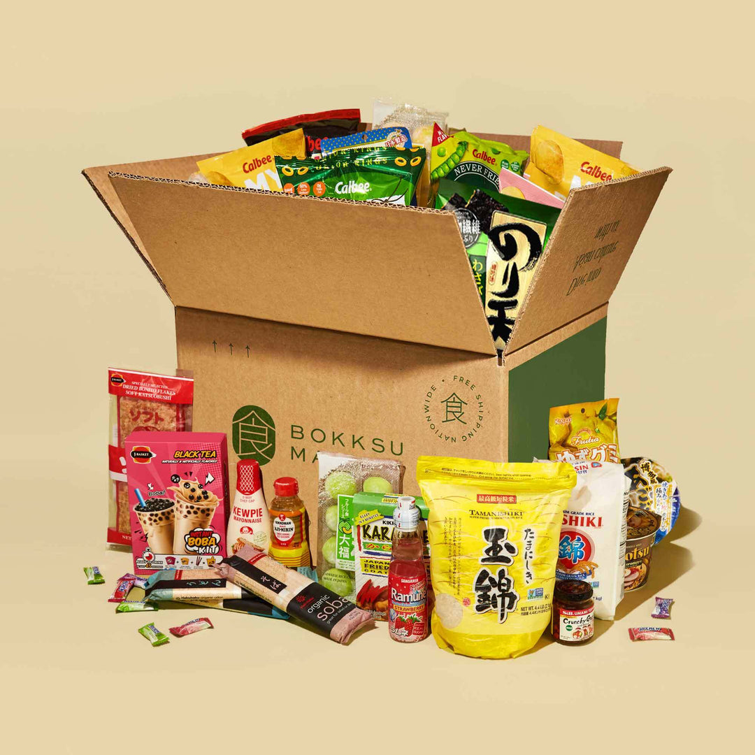 A large open cardboard box labeled "Bokksu Market," filled with various Japanese snacks, instant foods, and other delightful treats from the Discovery Pack. Additional products are displayed in front of the box, creating an enticing array of flavors.