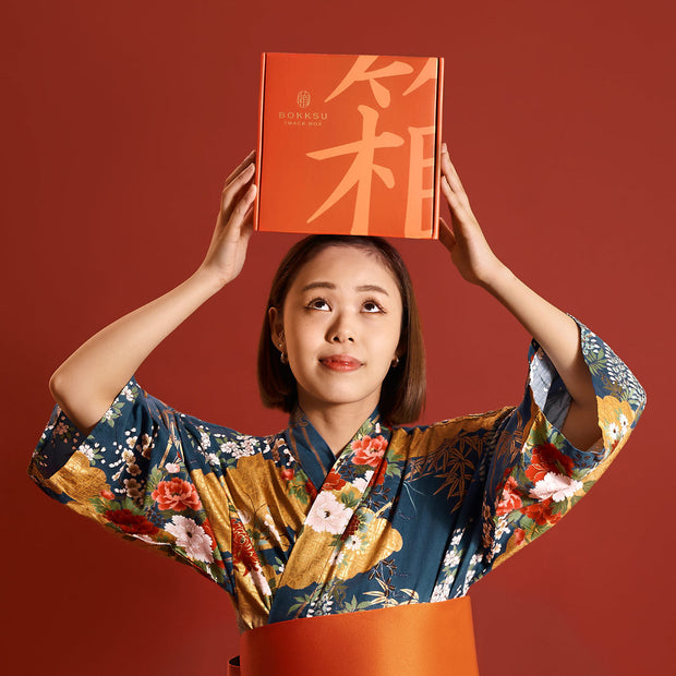 In front of an orange backdrop, a person in a floral kimono lifts the Bokksu Snack Box: Seasons of Japan (FBM) by Bokksu above their head.
