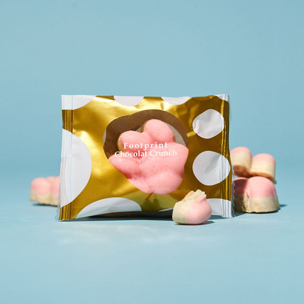 A gold and white polka dot package labeled "Footprint Chocolat Crunch" is displayed with pink and white chocolate pieces on a light blue background—a tasty snack from Bokksu’s The Cat Lover's Box.
