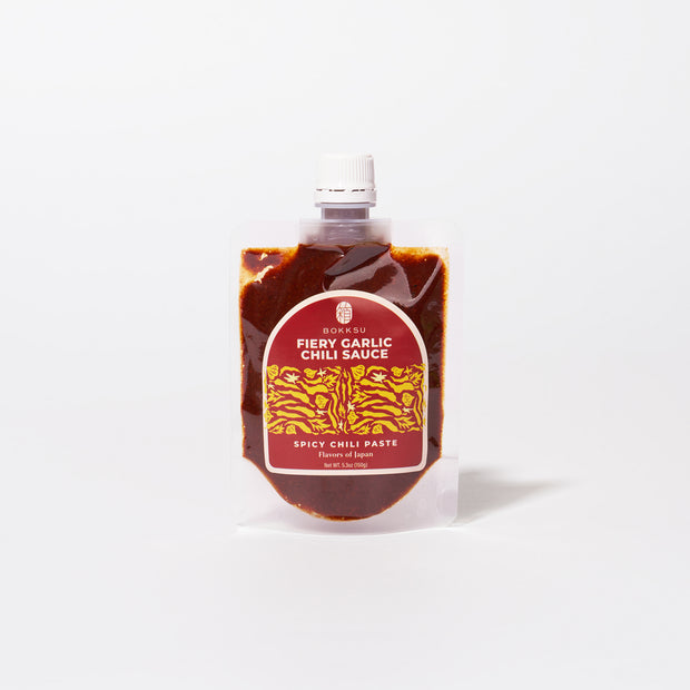 A pouch of Bokksu Fiery Garlic Chili Sauce, a Japanese chili garlic paste, with a white cap and red label featuring yellow accents, stands upright against a plain white background.