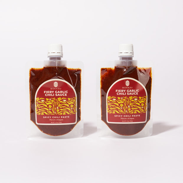 Two Bokksu Fiery Garlic Chili Sauce (2-Pack) pouches, featuring red labels and filled with dark spicy chili garlic paste, made in Japan, are shown side by side on a white background.