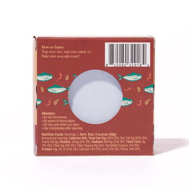 Back of the Bokksu Spicy Japanese Sardines package displays nutrition facts, instructions, a barcode, and a circular window revealing sardines in togarashi pepper. A fish pattern on a reddish-brown background adds charm.