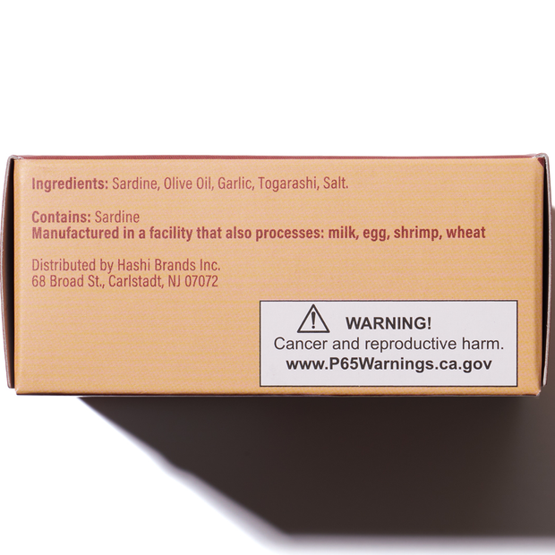 Side of Bokksu Spicy Japanese Sardines package listing ingredients like olive oil, iwashi, and togarashi pepper, allergen info, and a Proposition 65 warning about cancer/reproductive harm with a website for details.