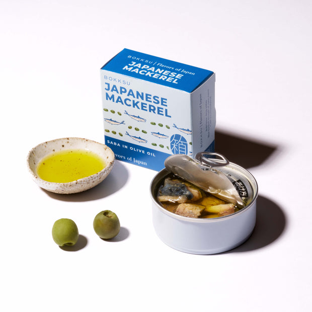 A can of Bokksu Japanese Mackerel in olive oil sits open beside its box, with a small bowl of olive oil and two green olives on a white surface.