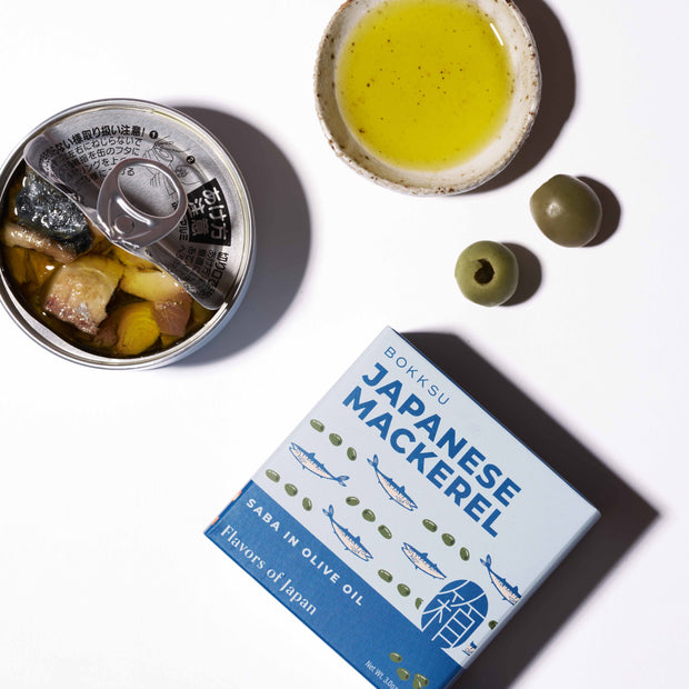 An opened can of Bokksu Japanese Mackerel in olive oil sits next to two green olives, a small dish of olive oil, and a box labeled "Japanese Mackerel" on a white surface.