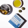 An opened can of Bokksu Japanese Mackerel in olive oil sits next to two green olives, a small dish of olive oil, and a box labeled "Japanese Mackerel" on a white surface. thumbnail 2