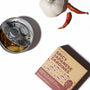 An open can of Bokksu Spicy Japanese Sardines in olive oil sits on a white surface with a garlic bulb, two dried red chili peppers, and its packaging—showcasing the bold togarashi pepper flavor. thumbnail 3