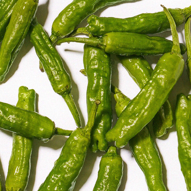 A top-down image of  peppers scattered on a white background, emphasizing their fresh texture, curved shapes, and vibrant green color, just like the ones used in Bokksu Japanese Yuzu Hot Sauce
