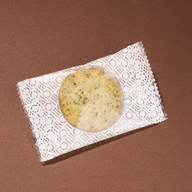 A single April '26 Bokksu cookie with dark specks, handpicked in Japan, is wrapped in a transparent, patterned plastic and set on a brown surface.