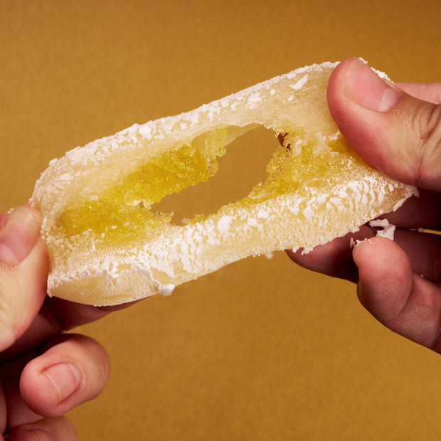 Two hands pull apart a translucent, yellow-filled, powdered candy from the April '26 Bokksu: Handpicked in Japan box, highlighting a unique regional specialty against a plain brown background.