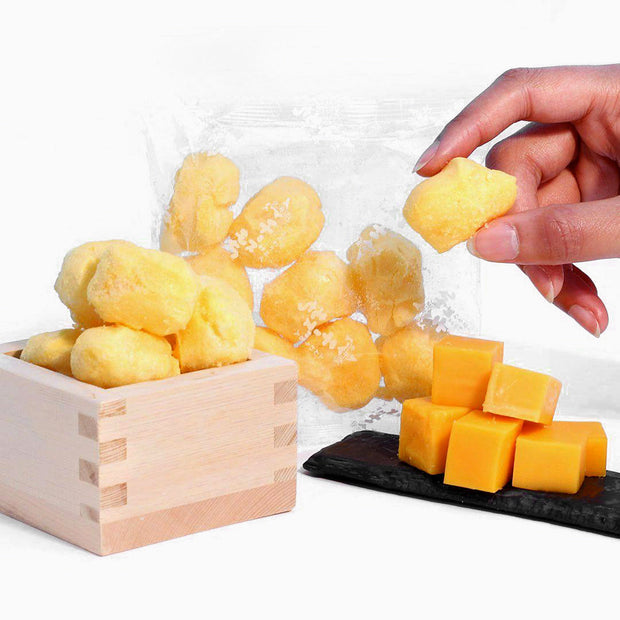 A hand picks up a yellow cheese puff from a wooden box, with more Doki Doki Snack Mix: Japan Mochi Puffs in a clear bag and cubes of cheese on a black tray nearby.