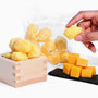 A hand picks up a yellow cheese puff from a wooden box, with more Doki Doki Snack Mix: Japan Mochi Puffs in a clear bag and cubes of cheese on a black tray nearby. thumbnail 10