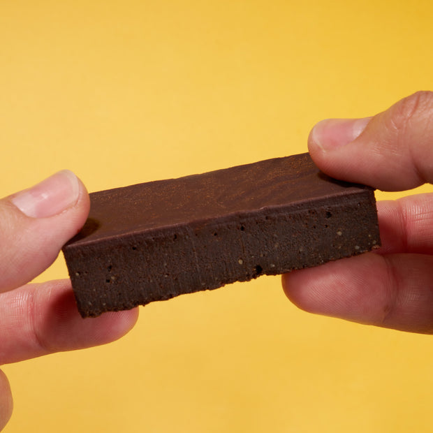 A person holds a rectangular chocolate brownie with both hands against a yellow background, evoking the treats from April '26 Bokksu: Handpicked in Japan.