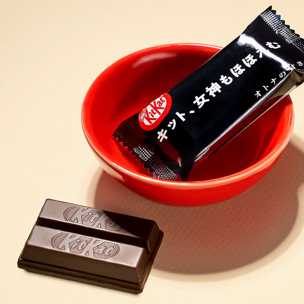 A piece of chocolate from the Dagashi Snack Crate - 30-Piece Japanese Snack Variety Box with Candy, Chocolate, Cookies, Mochi & Ramune Treats sits wrapped in black packaging in a red bowl next to an unwrapped Kit Kat on a beige surface.