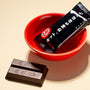 A piece of chocolate from the Dagashi Snack Crate - 30-Piece Japanese Snack Variety Box with Candy, Chocolate, Cookies, Mochi &amp; Ramune Treats sits wrapped in black packaging in a red bowl next to an unwrapped Kit Kat on a beige surface. thumbnail 27