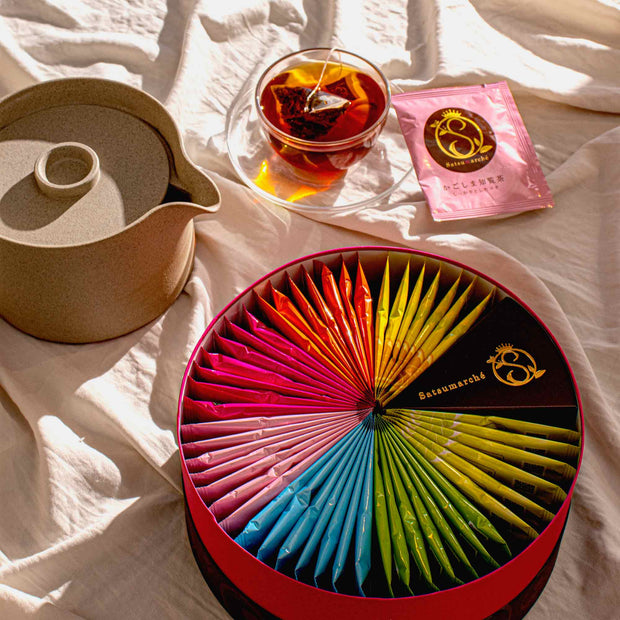 A Kagoshima Seicha Gift Box: Satsumarche Tea Bags (49 Pieces, 7 Flavors) is displayed with a teapot and a glass cup of tea with a teabag, set on a white cloth background.