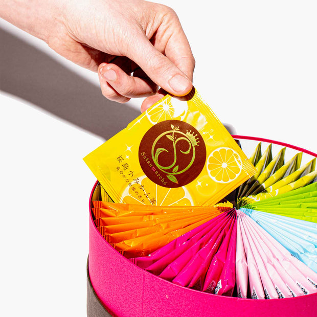 A hand picks up a yellow Satsumarche tea bag from the Kagoshima Seicha Gift Box: Satsumarche Tea Bags (49 Pieces, 7 Flavors), which features colorful assorted packets arranged in a fan shape.
